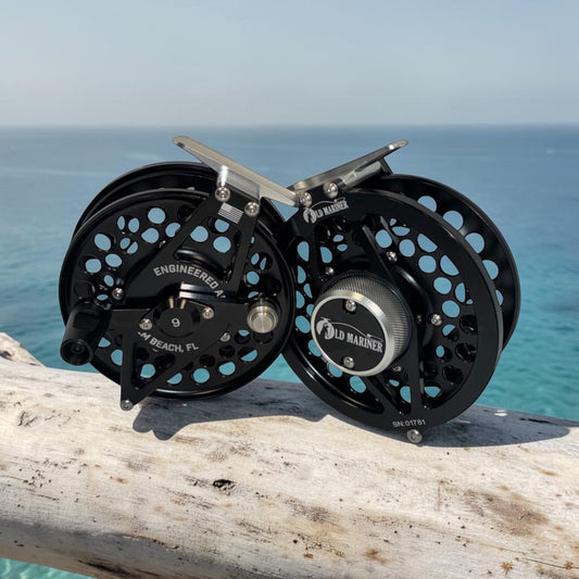 Two black Old Mariner fly fishing reels resting on a weathered wooden log with the ocean in the background.